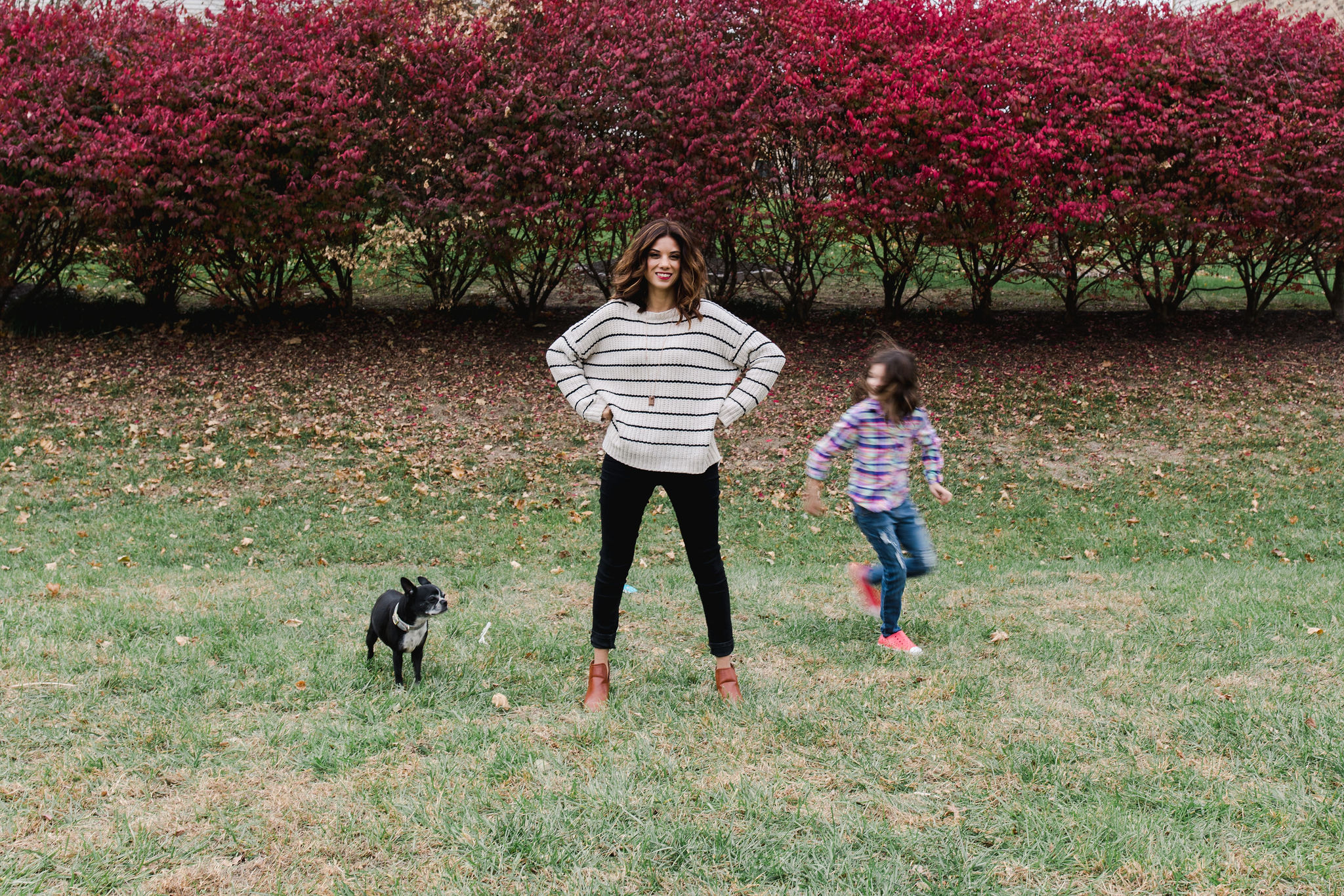 Illiah, a woman dressed in black and white, stands in front of a row of bright azalea bushes with her dog and daughter running around her, out of focus